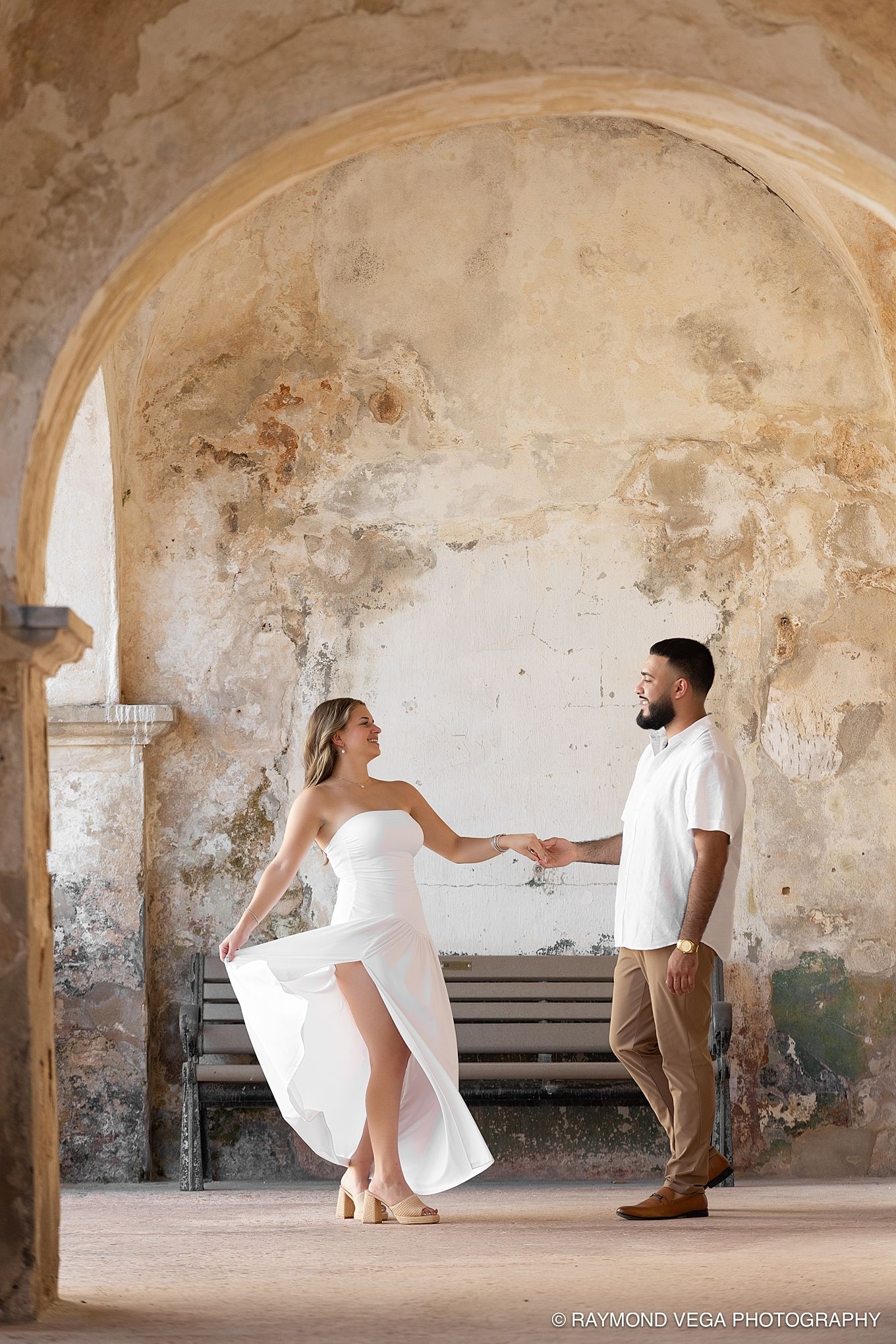 Couple dancing under stone arch in white outfits at historic venue, engagement photo by Raymond Vega Photography. Castillo de San Cristóbal