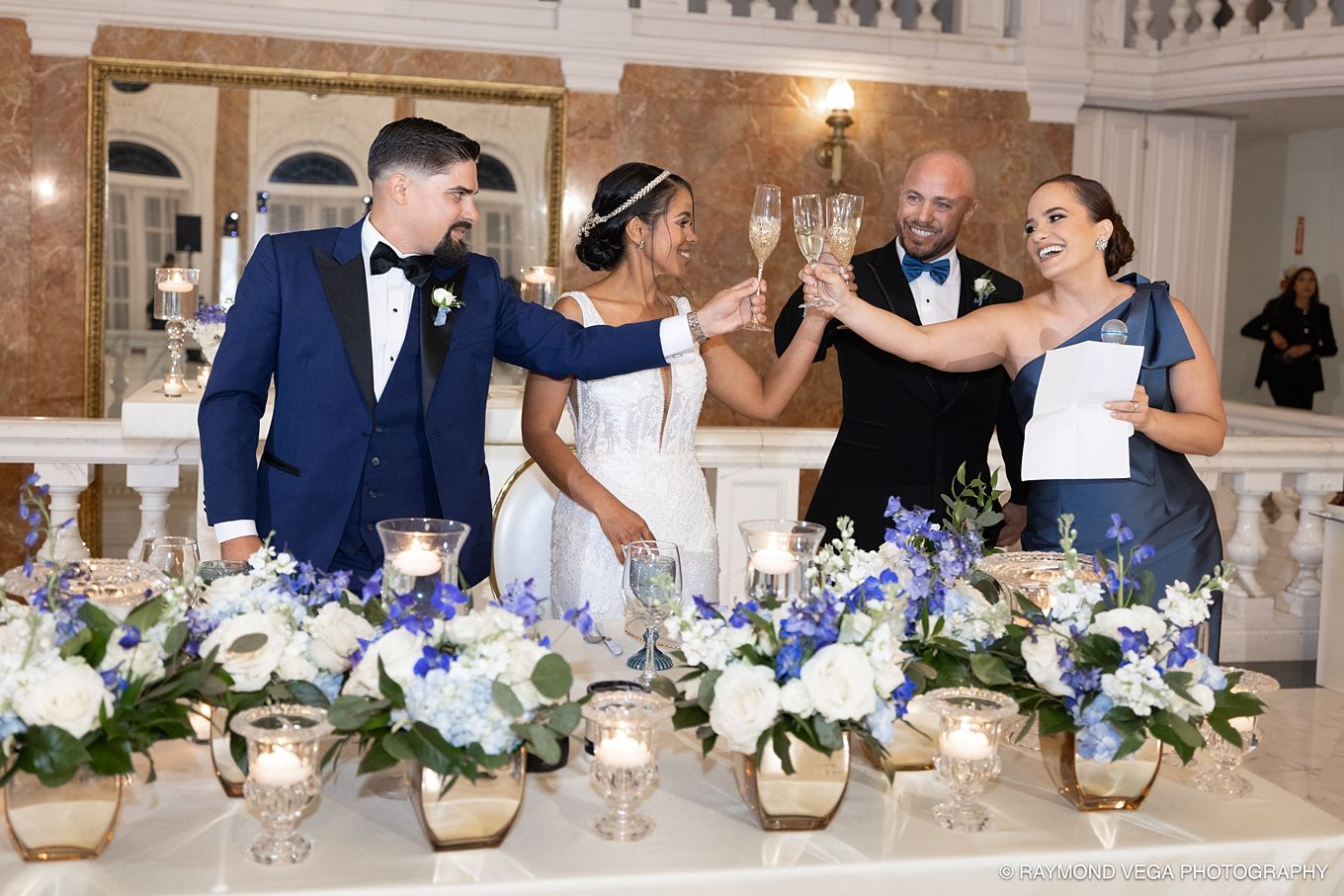 Wedding toast with bride and groom in blue tuxedo clinking champagne glasses with guests at elegant floral reception table.