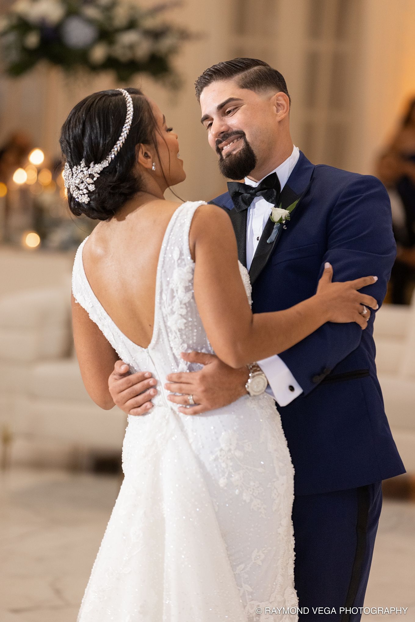 Bride and groom share first dance, she wears backless lace gown with hair piece, he wears navy tuxedo.