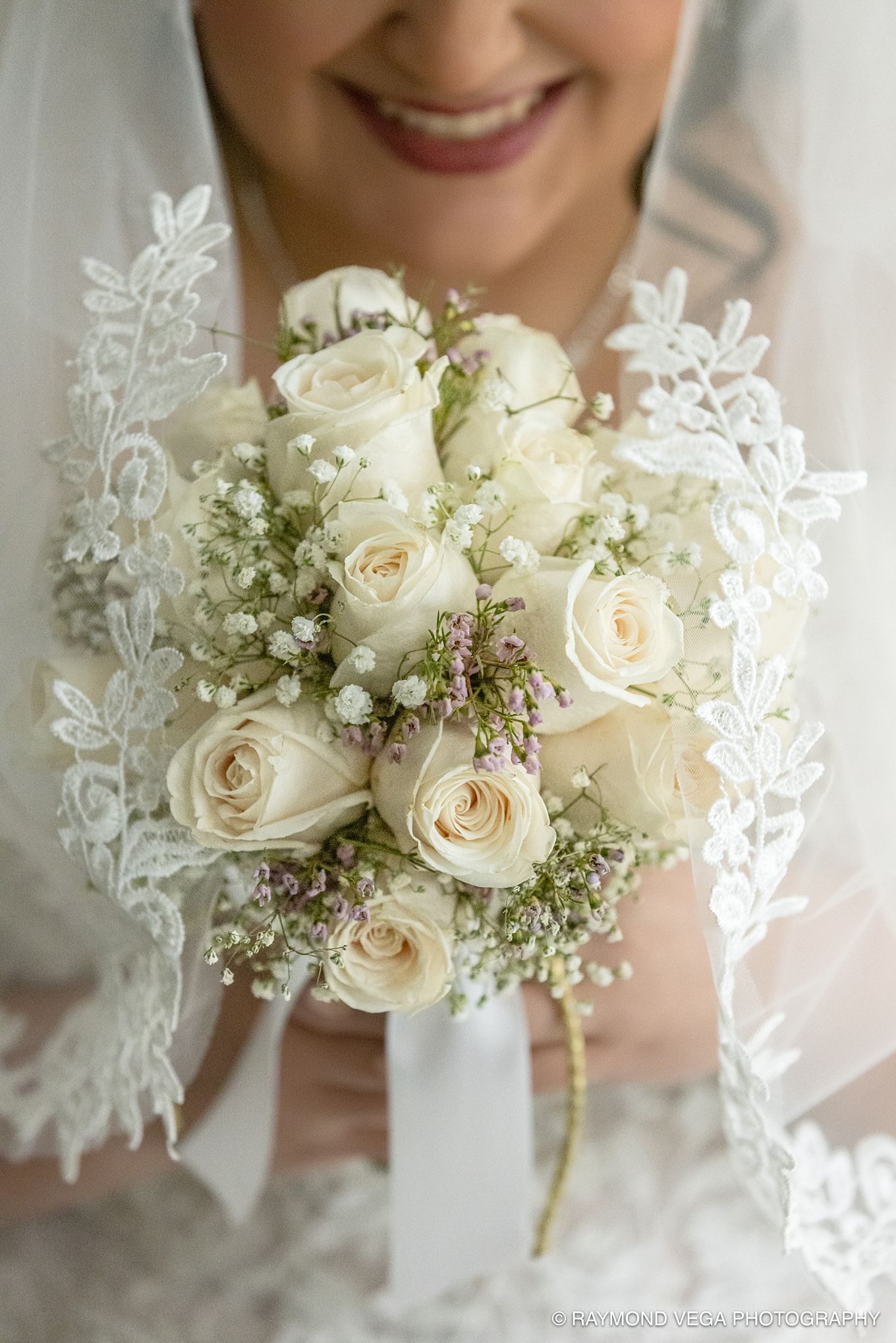 Smiling bride in lace veil holding elegant white rose and baby's breath wedding bouquet with purple accents.