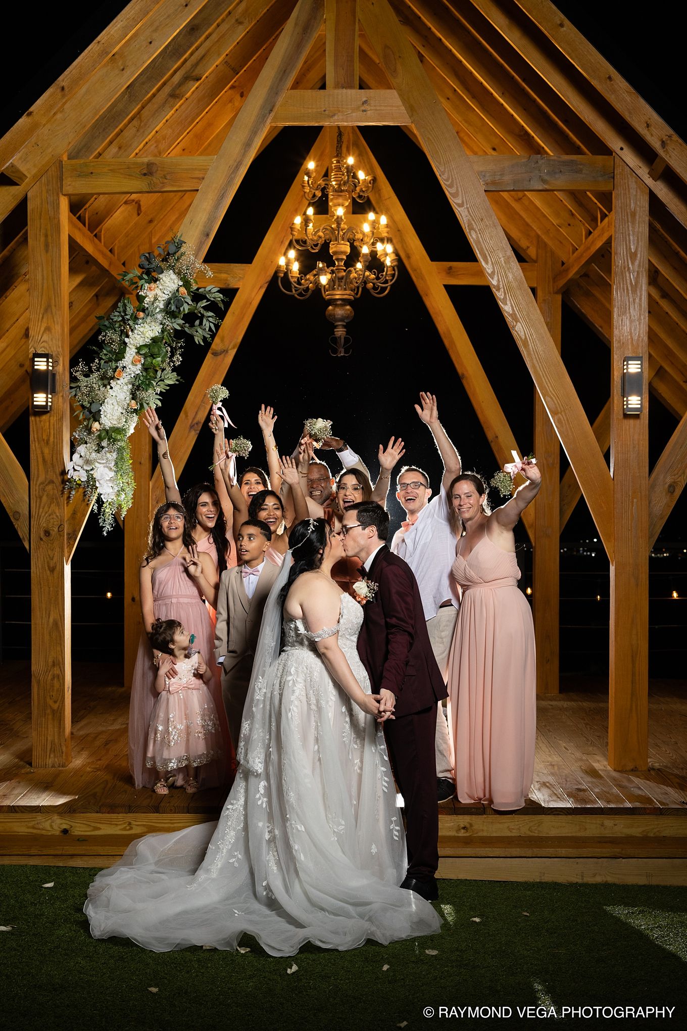 Wedding party celebrating under a wooden arch with chandelier at night, bride and groom kissing surrounded by cheering bridesmaids.