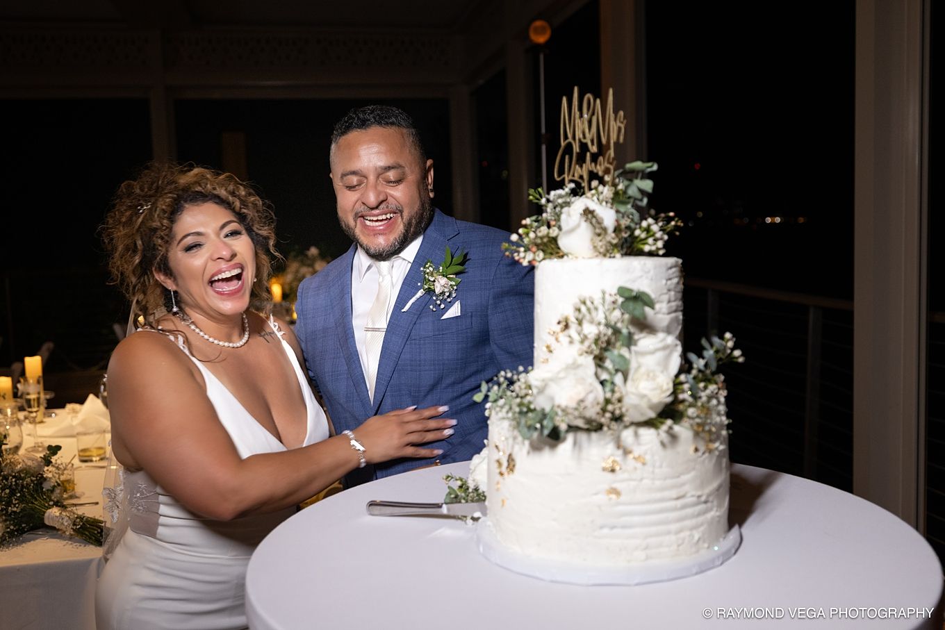 Joyful couple cutting their elegant three-tier white wedding cake adorned with greenery and florals at their reception.