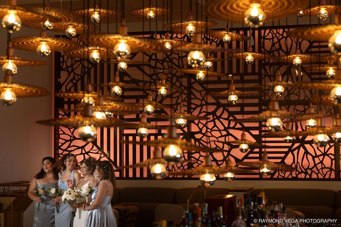 Bridesmaids in gray dresses stand beneath warm glowing pendant lights in front of an ornate decorative wall at a wedding venue.