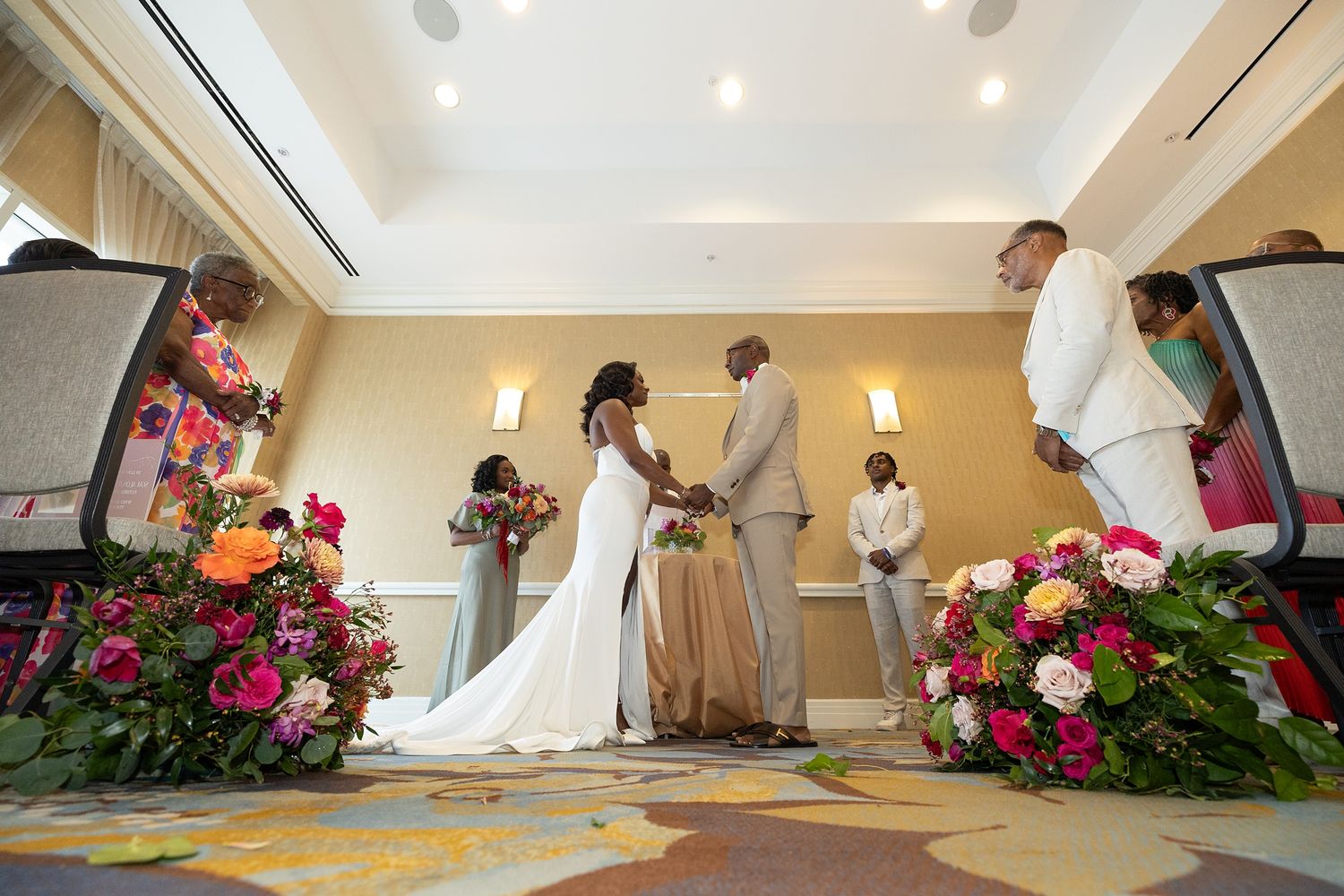 Black couple exchanges vows at elegant indoor wedding ceremony surrounded by vibrant floral arrangements and guests. — Raymond Vega