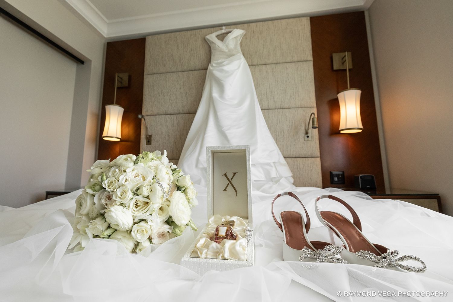 Elegant wedding dress hanging in hotel room with bridal bouquet of white roses and silver heels on white bed. Hotel Condado Vanderbilt
