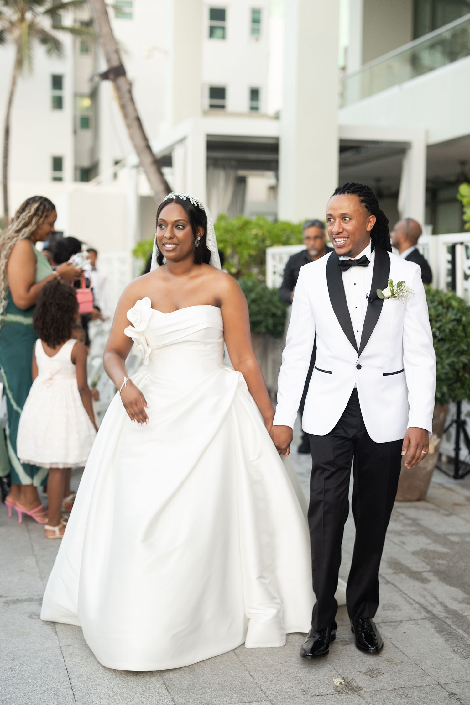 Bride in strapless white ball gown and groom in white tuxedo jacket walk hand in hand at outdoor wedding venue. Hotel La Concha Puerto Rico