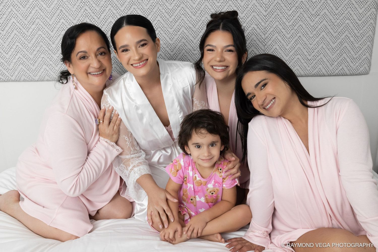 Five women and a young girl in pink robes and pajamas pose together on a bed for a cheerful family photo.
