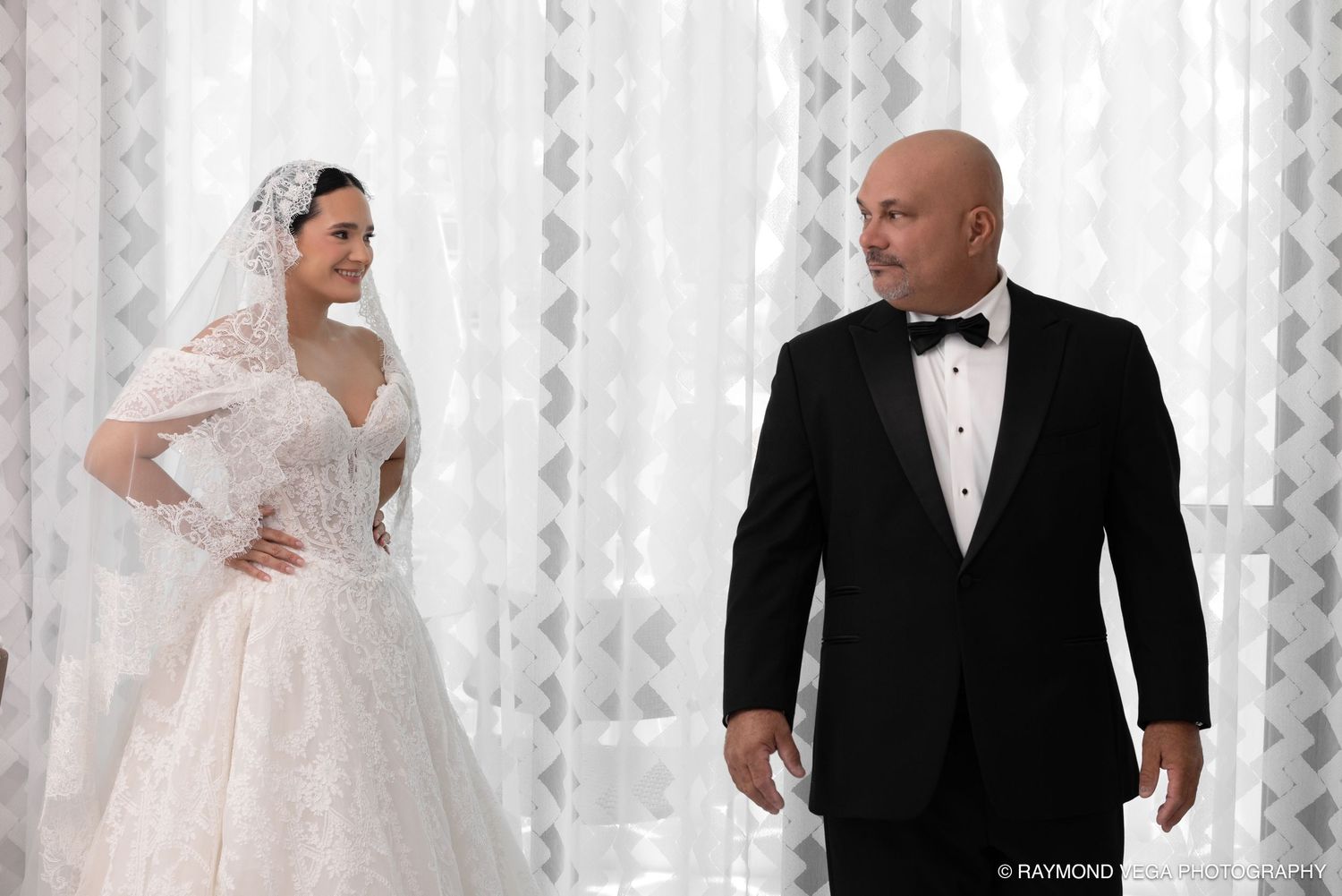 Bride in lace wedding gown with veil stands beside groom in black tuxedo at elegant wedding venue.