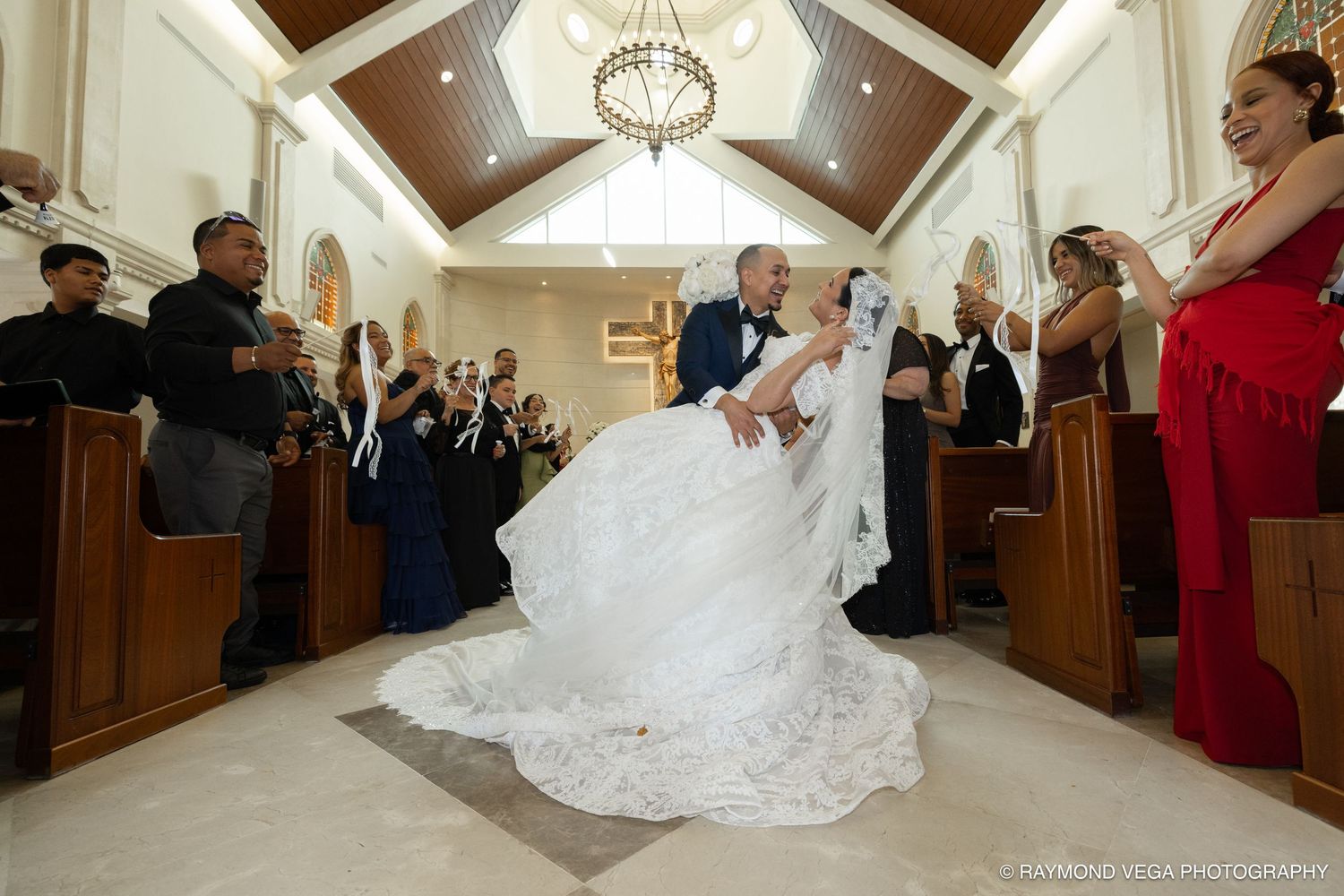 Groom dips bride in stunning white gown during wedding ceremony in elegant chapel with guests applauding.