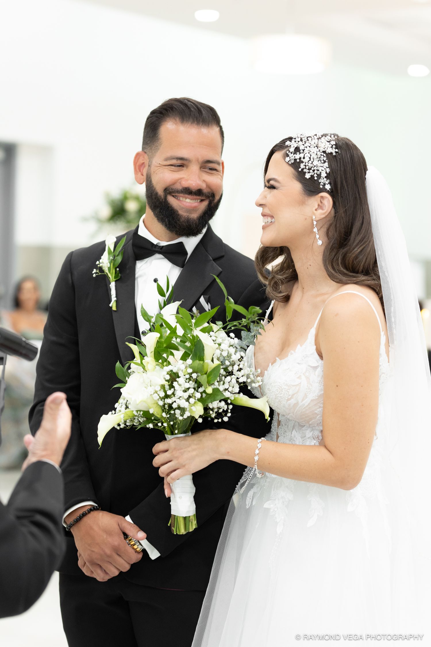 Smiling bride and groom exchange vows at wedding ceremony, bride holds white floral bouquet with greenery.