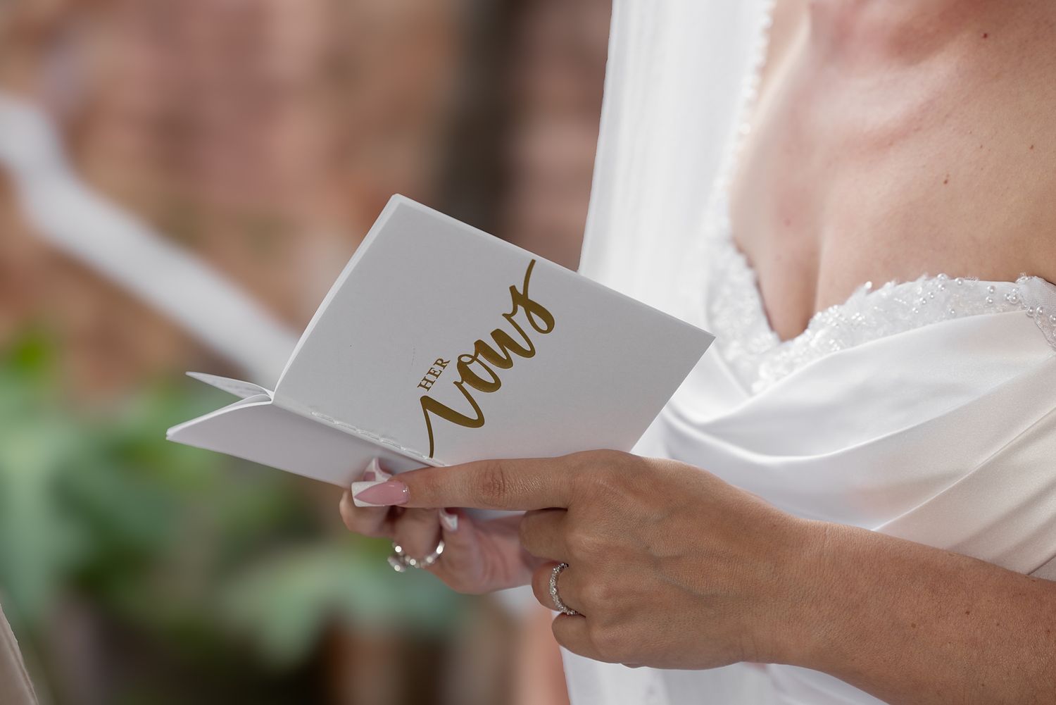 Bride in white lace dress holding a gold and white wedding vows booklet during an outdoor ceremony.