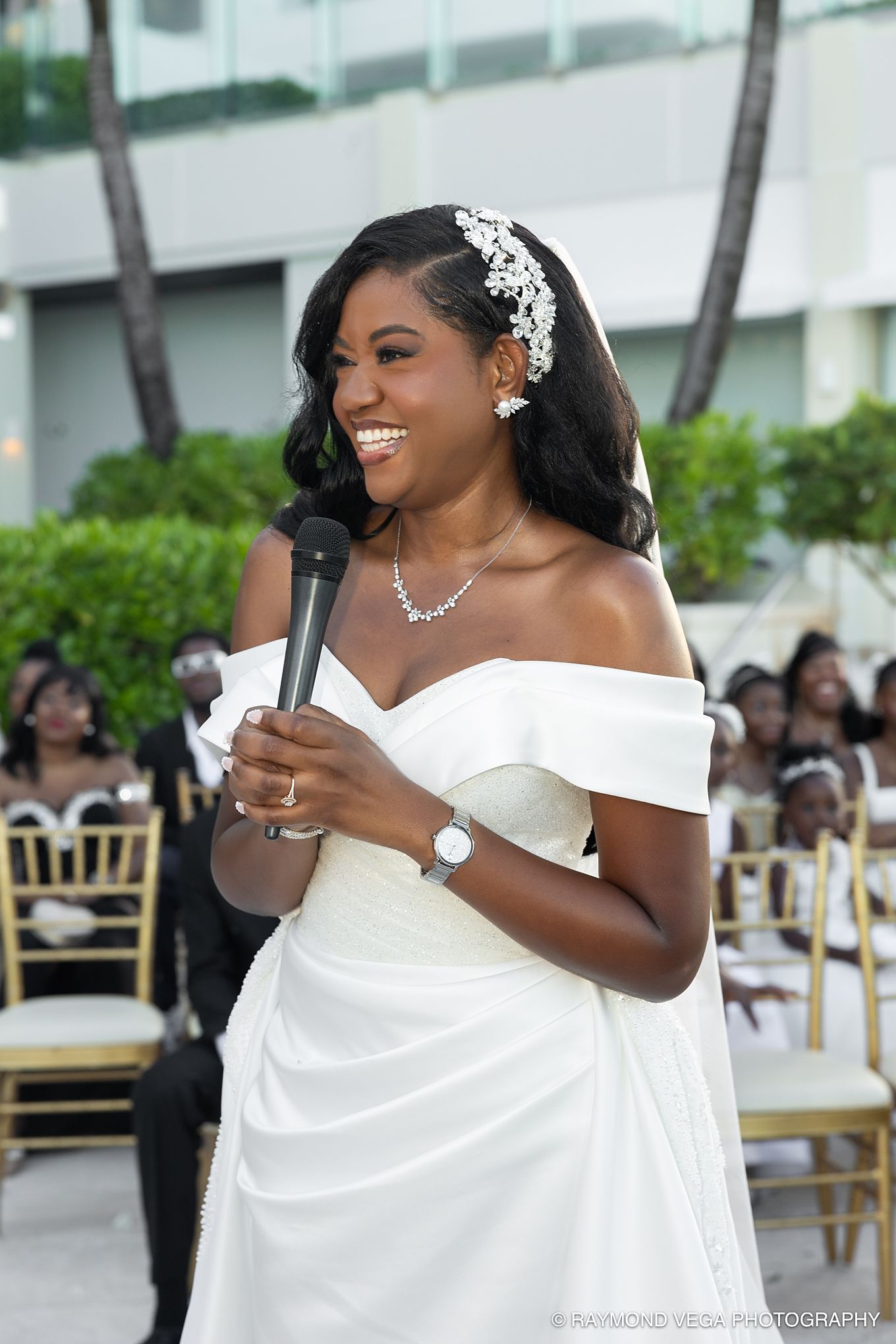 Smiling bride in off-shoulder white gown holds microphone at outdoor wedding ceremony, wearing pearl headpiece and necklace.