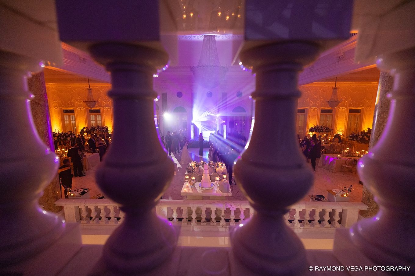Elegant wedding reception venue photographed through white balusters with purple lighting and guests dancing on the floor below.