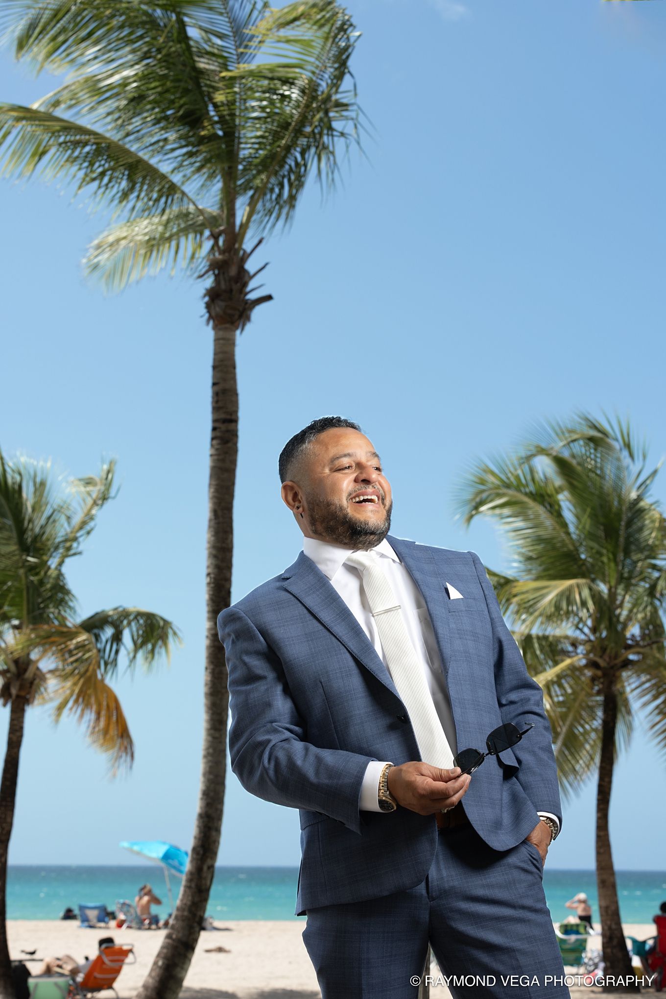 Man in blue suit holding sunglasses smiles on tropical beach with palm trees and ocean backdrop on sunny day.