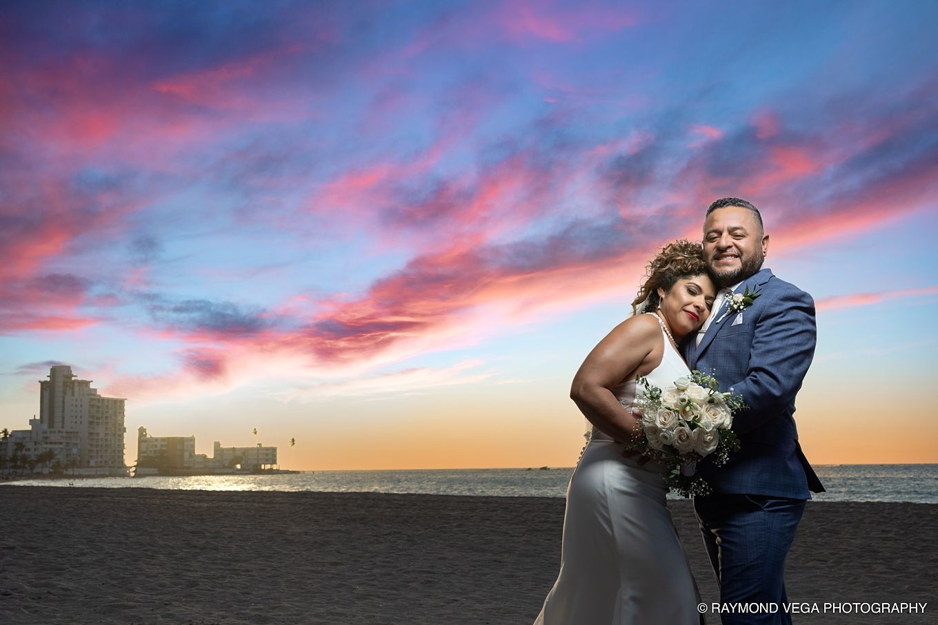 Bride and groom embrace on beach at sunset with vibrant pink and blue sky backdrop in romantic wedding portrait. at Courtyard by Marriott