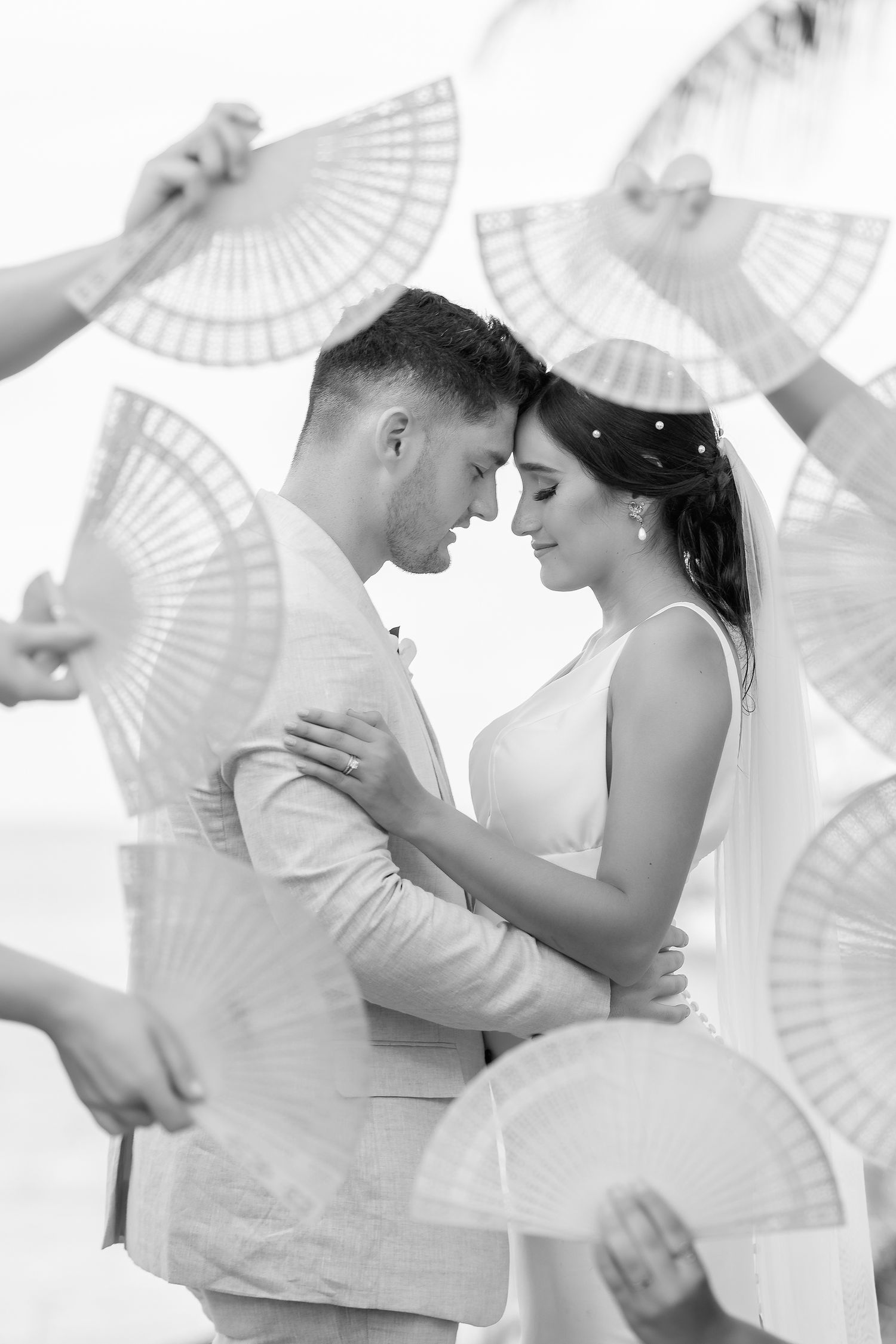 Black and white wedding photo of bride and groom embracing, surrounded by hands holding decorative fans. Raymond Vega Photography & Cinema
