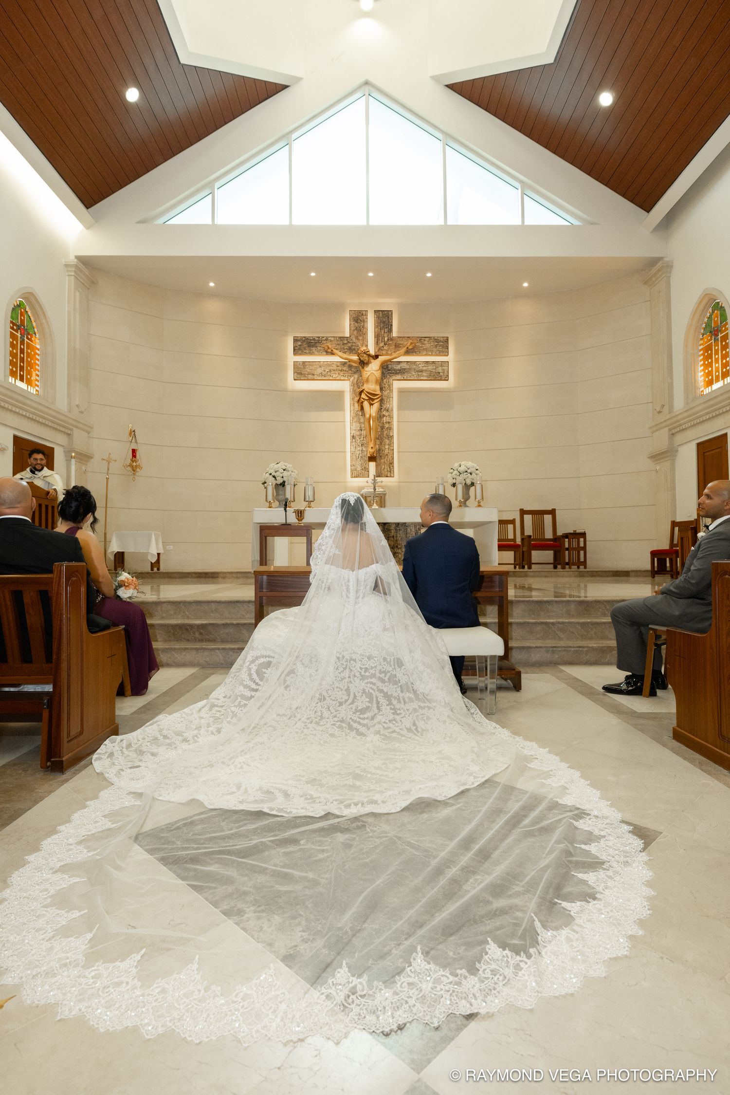 Bride and groom kneel at altar during Catholic wedding ceremony, bride's long lace cathedral veil trailing down church aisle.