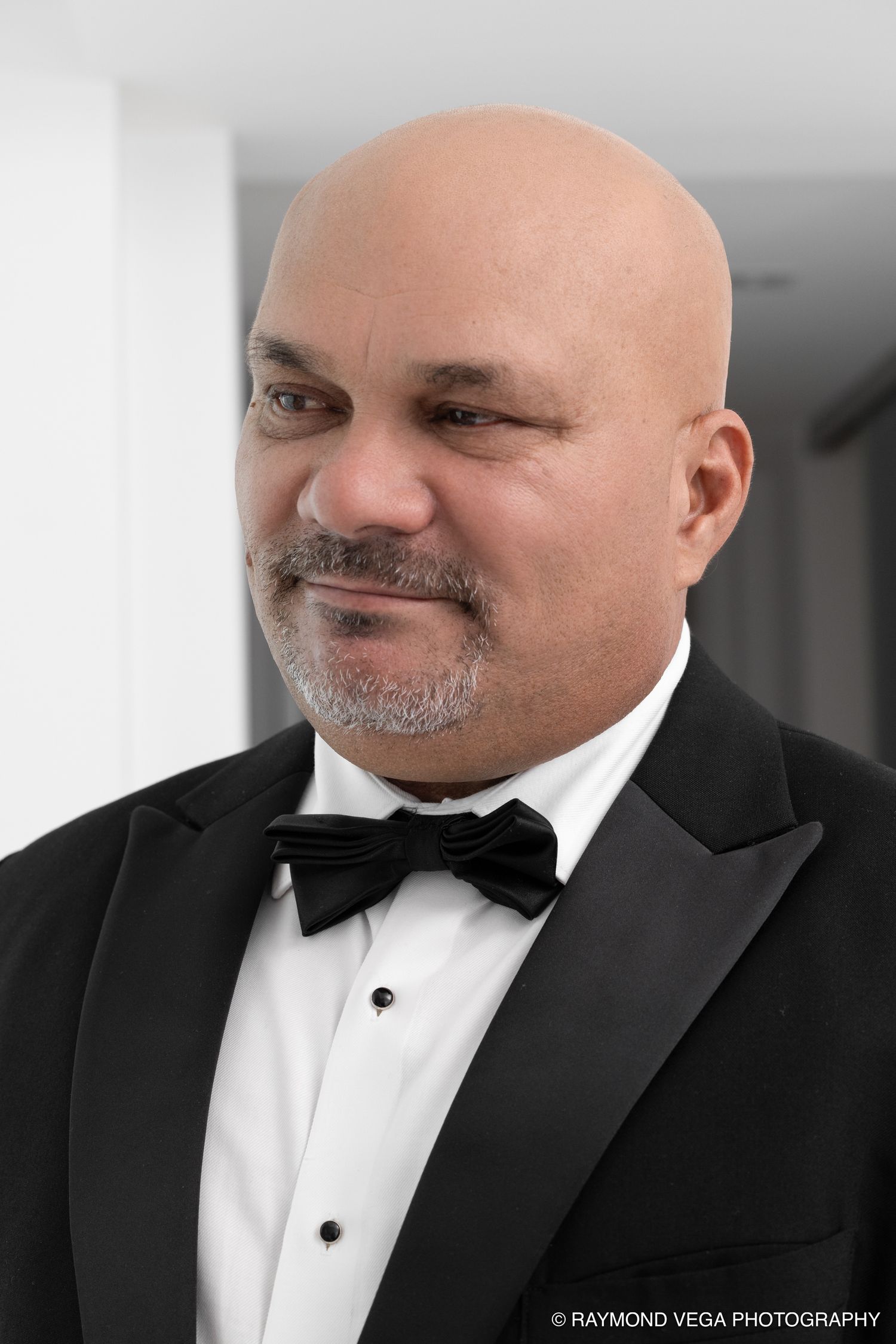 Bald man with goatee wearing black tuxedo and bow tie, smiling in professional portrait photo.