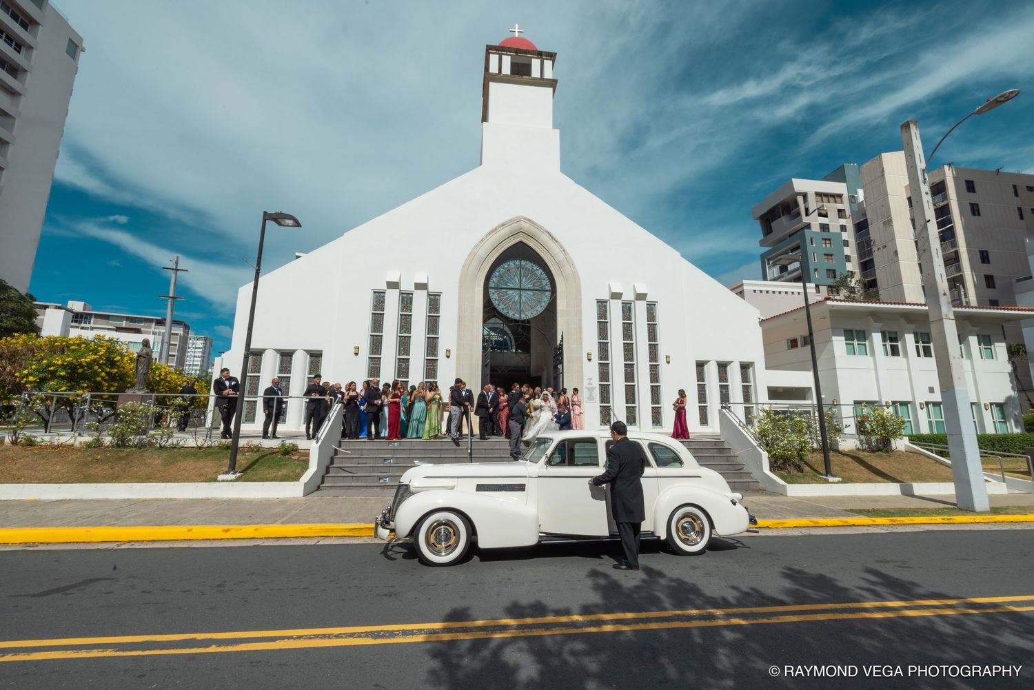 Bride and groom pose beside a vintage white car outside a modern white church on their wedding day.