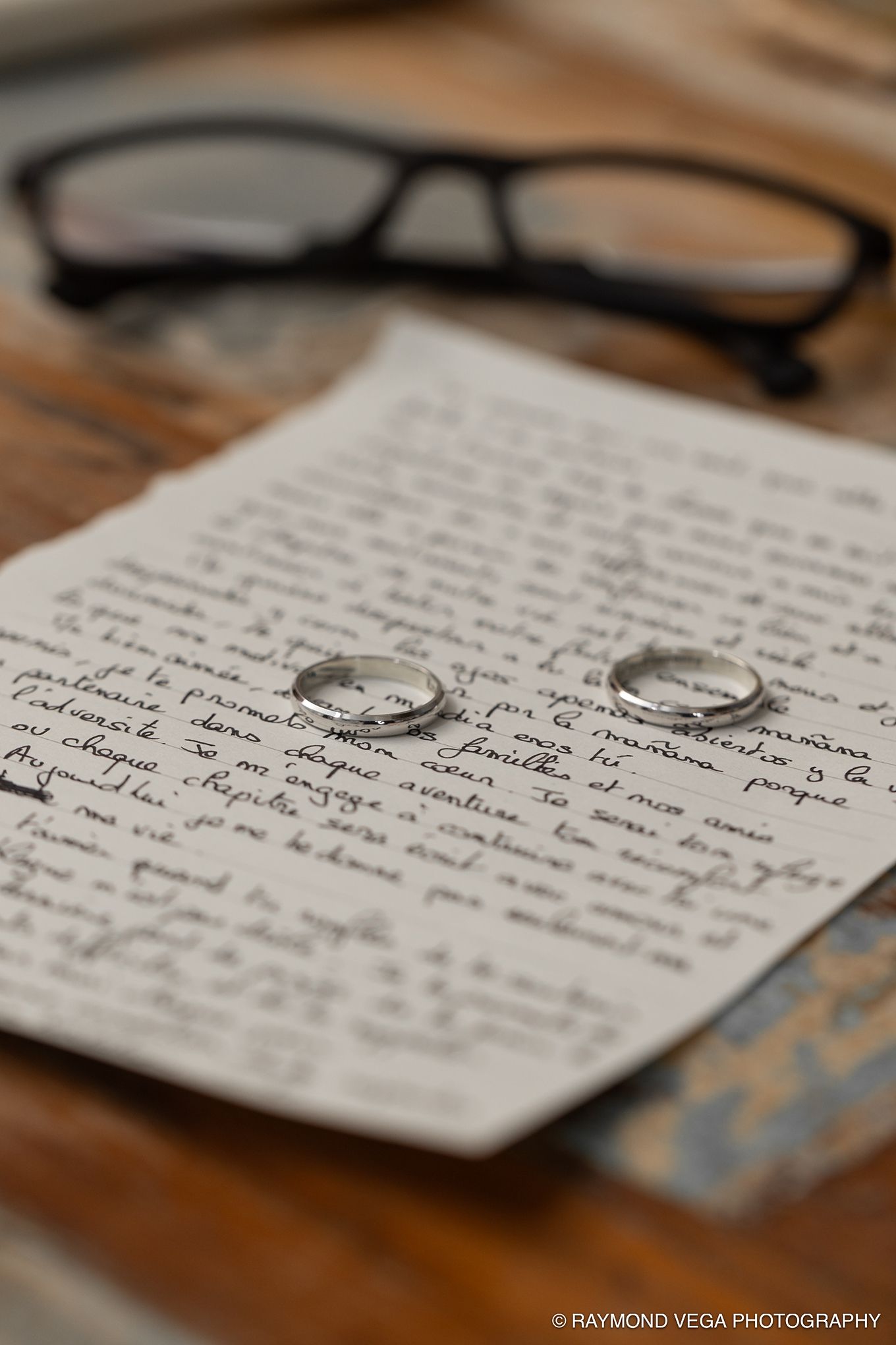 Two silver wedding bands rest on a handwritten love letter beside reading glasses on a wooden table.