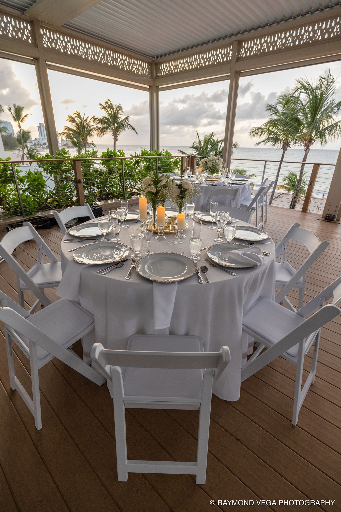 Elegant outdoor beachfront wedding reception setup with white round tables, chairs, and ocean views at sunset at Courtyard by Marriott