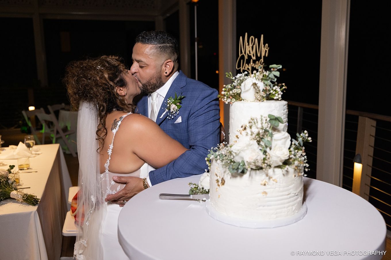 Bride and groom sharing a kiss beside a white floral wedding cake at an elegant outdoor evening reception at Courtyard by Marriott