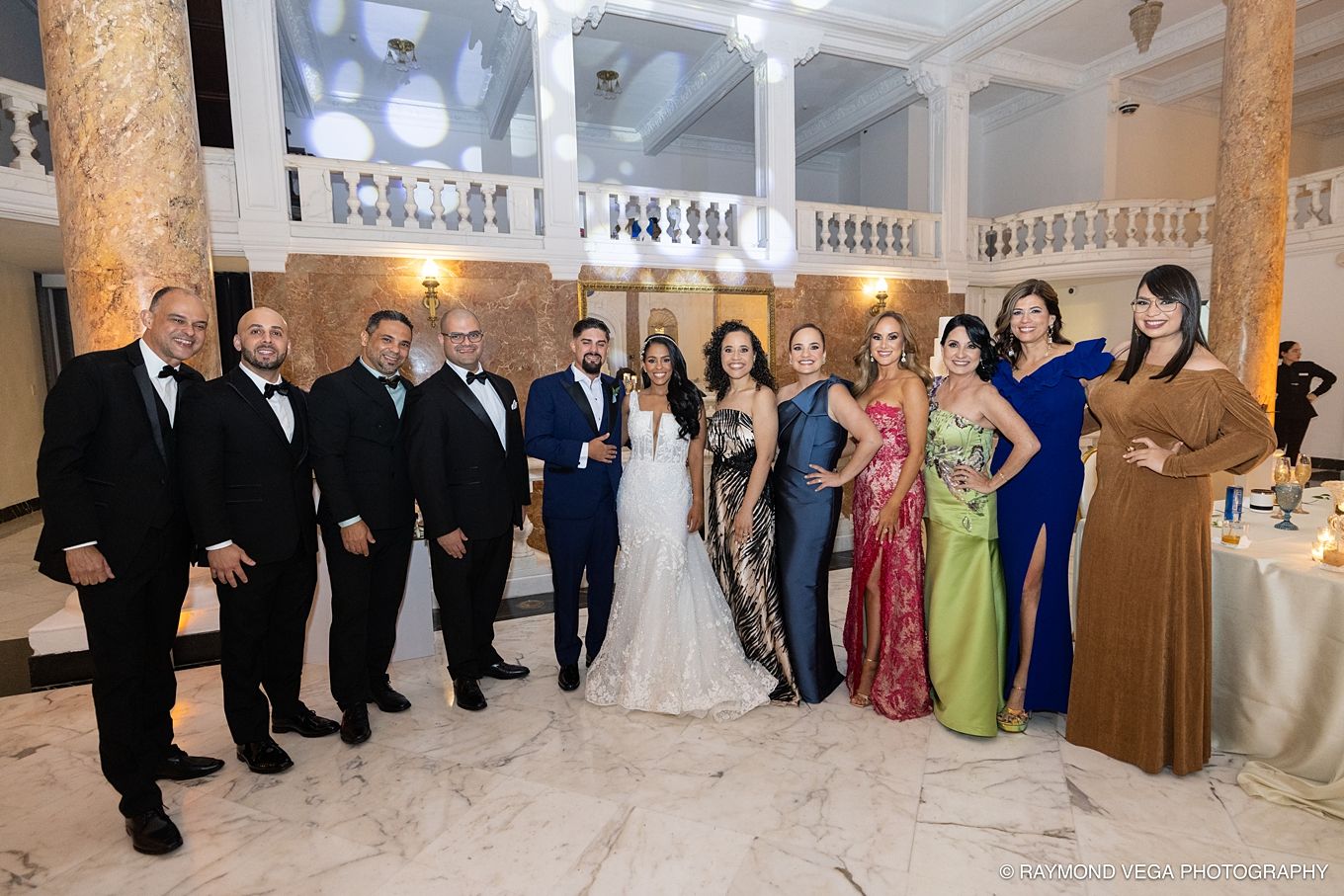 Wedding party poses in elegant marble venue, bride in white gown with groom and guests in formal attire.