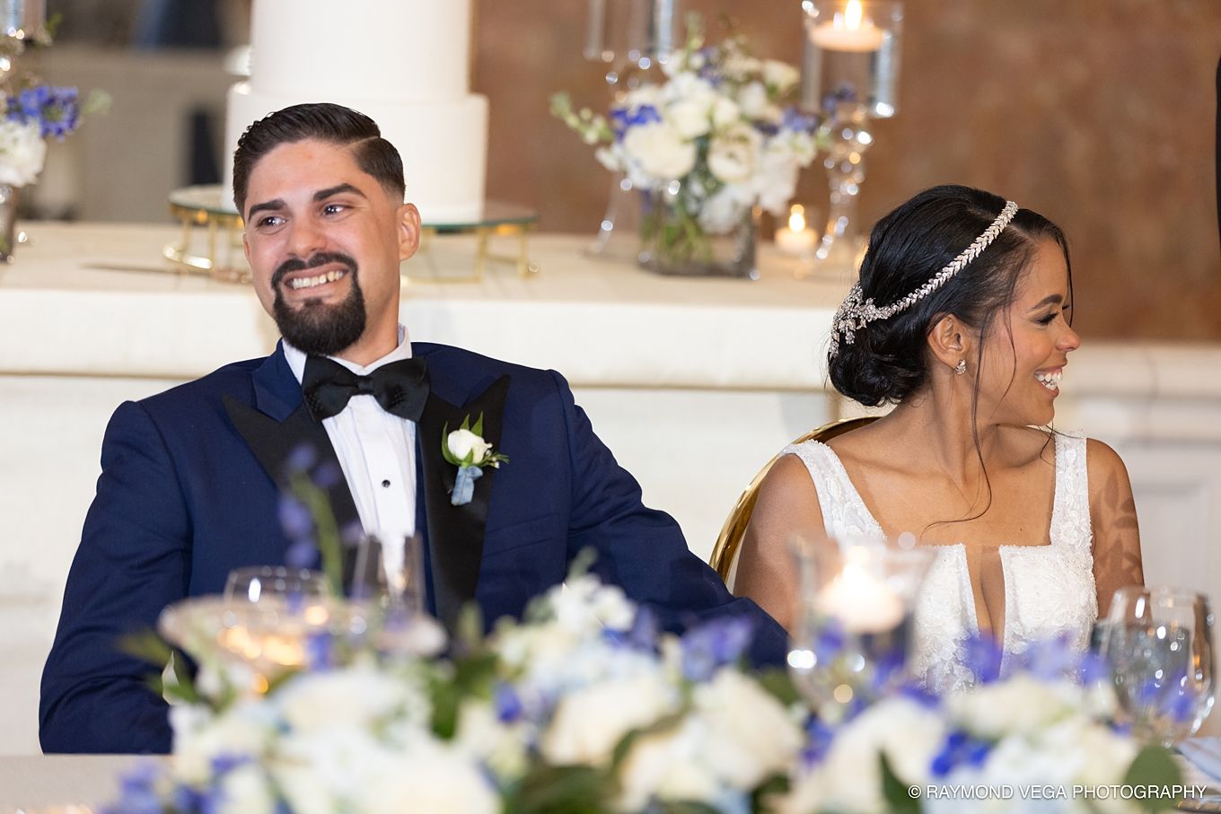 Smiling groom in navy tuxedo and bride in white gown with headpiece seated at wedding reception table with blue floral arrangements.
