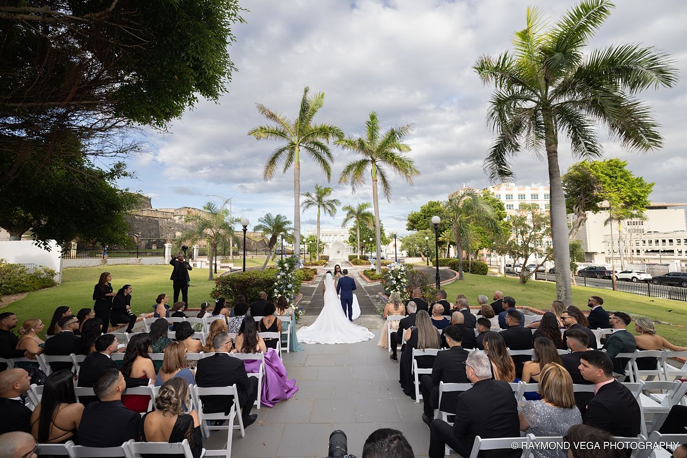 Outdoor wedding ceremony with guests seated on white chairs along a palm tree-lined aisle under a cloudy sky. Antiguo Casino Puerto Rico