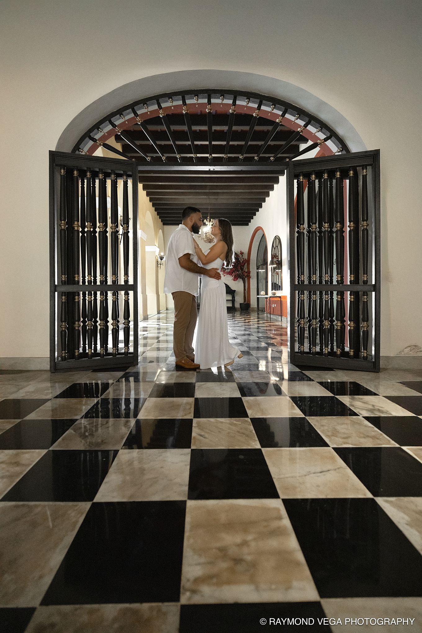 Couple embracing under arched doorway on checkered marble floor in elegant colonial building, captured by Raymond Vega Photography.