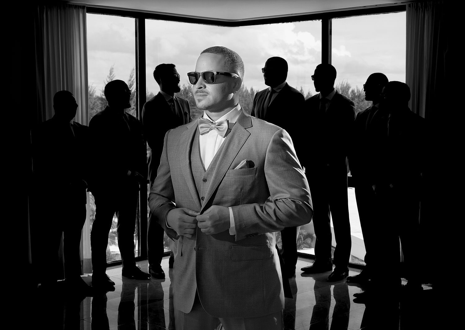 Black and white photo of a stylish groom in a suit and bow tie with groomsmen silhouetted behind him. Raymond Vega Photography & Cinema