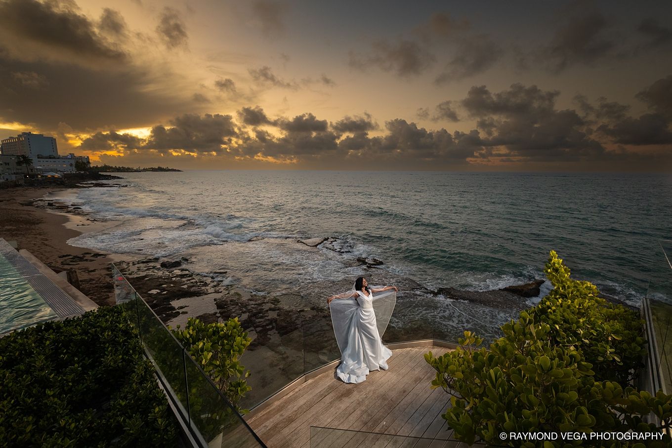 Bride in white gown stands on coastal terrace at sunset with dramatic stormy skies over the Caribbean sea.
