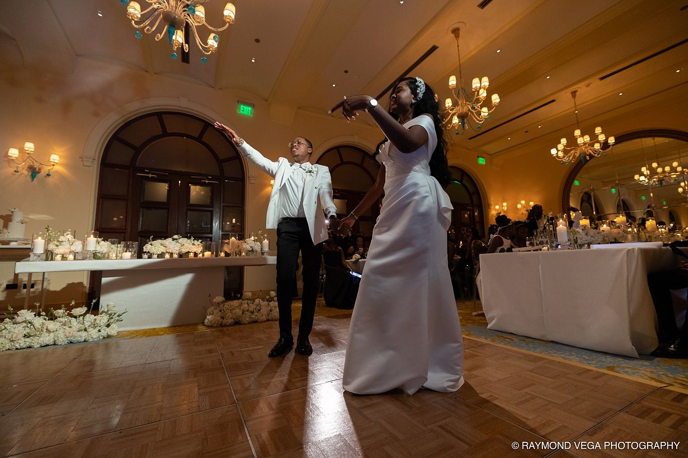 Bride in white gown plays violin while groom gestures during elegant wedding reception in chandelier-lit ballroom.