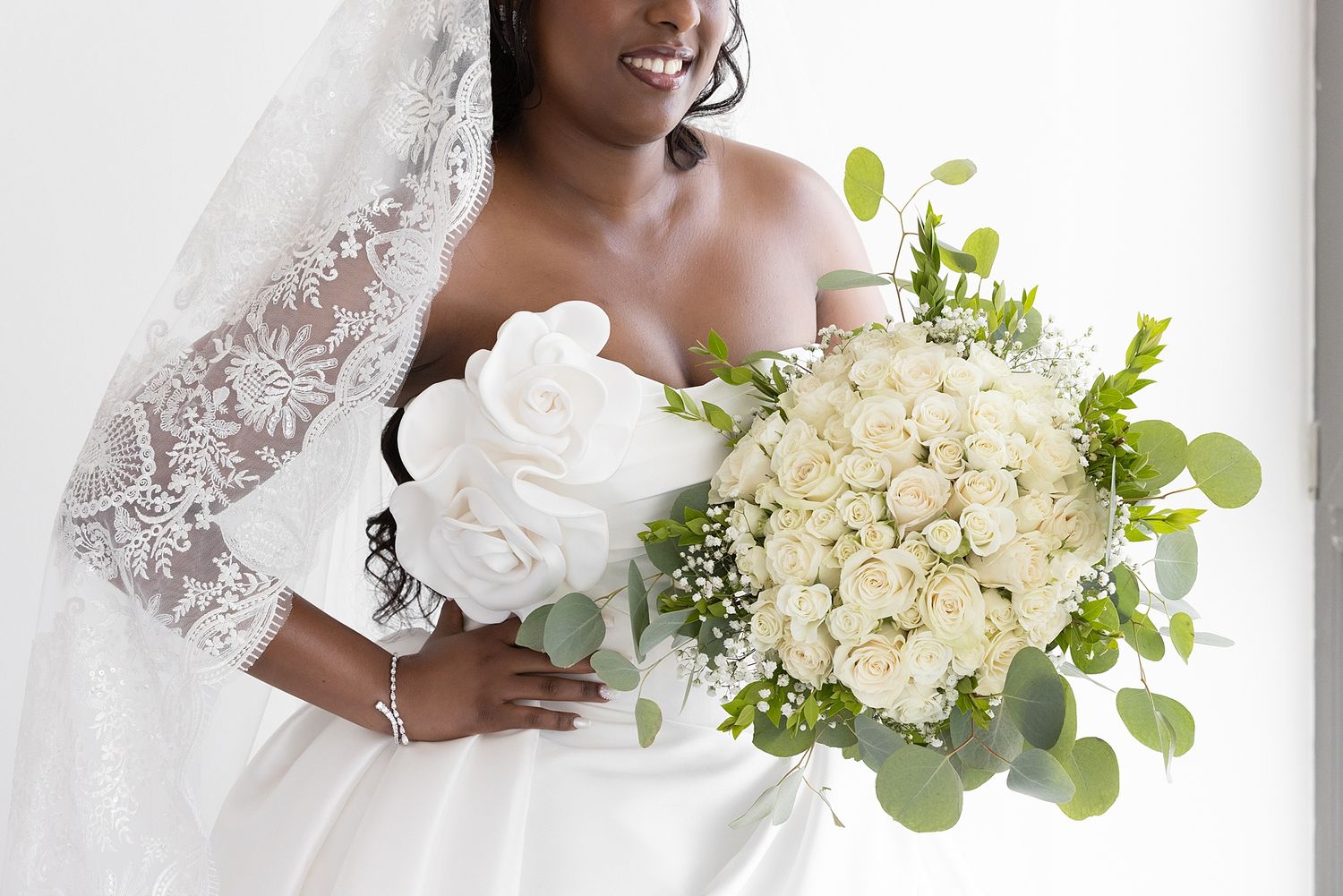 Bride in white lace wedding dress holding large bouquet of white roses and greenery, smiling elegantly.