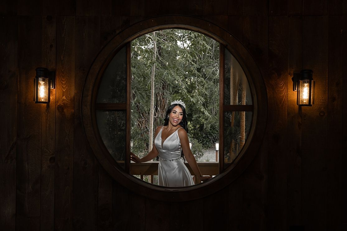 An elegant bride stands framed in a circular porthole window flanked by warm lanterns in a rustic wooden setting.