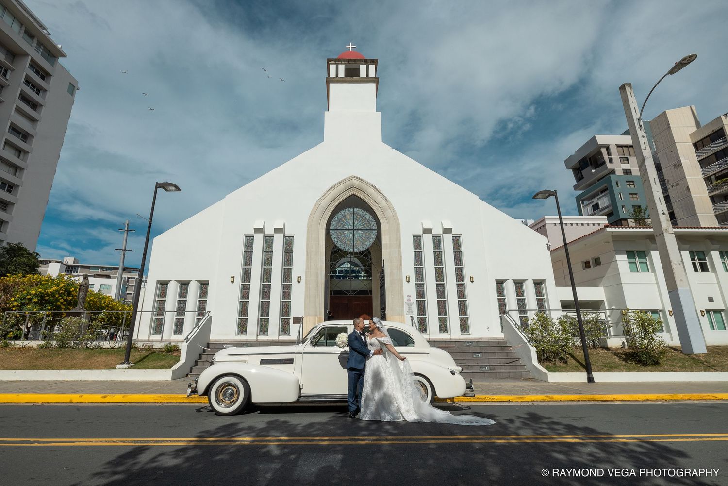 Bride and groom pose with vintage white car in front of a white church on their wedding day.