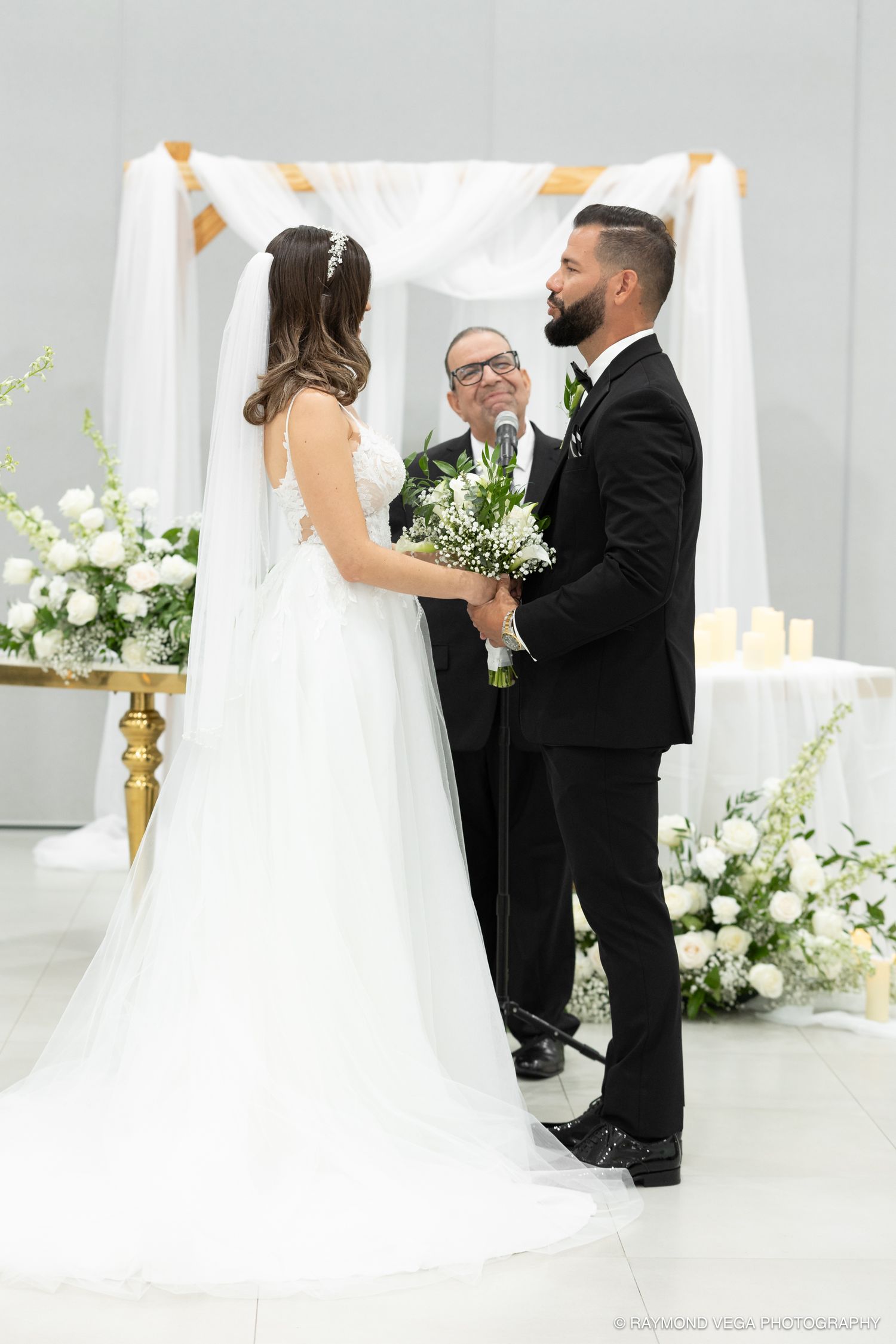 Bride and groom holding hands at elegant white floral wedding ceremony altar with officiant and draped canopy backdrop.