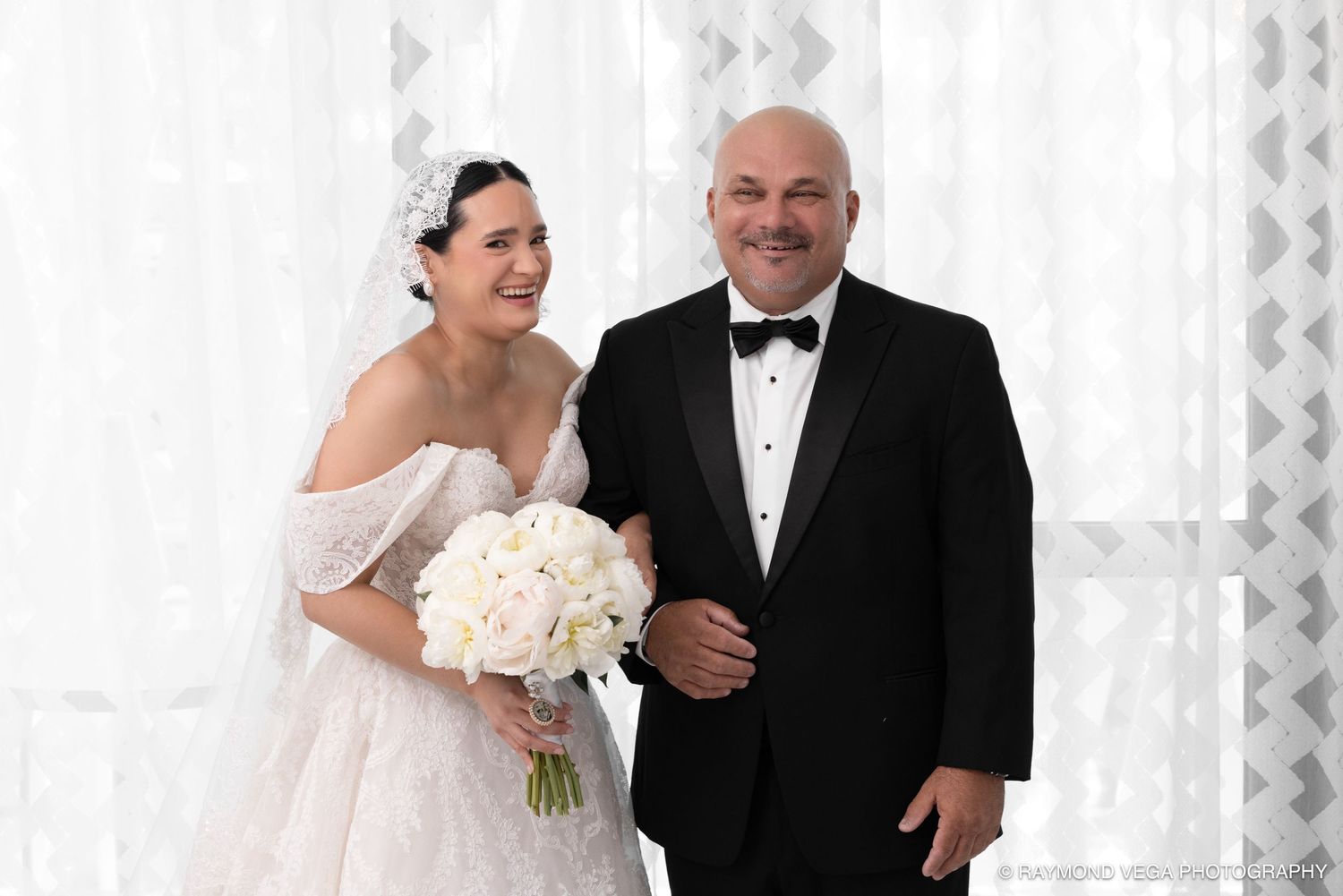 Bride in white off-shoulder gown holding bouquet stands smiling with man in tuxedo at elegant wedding venue.