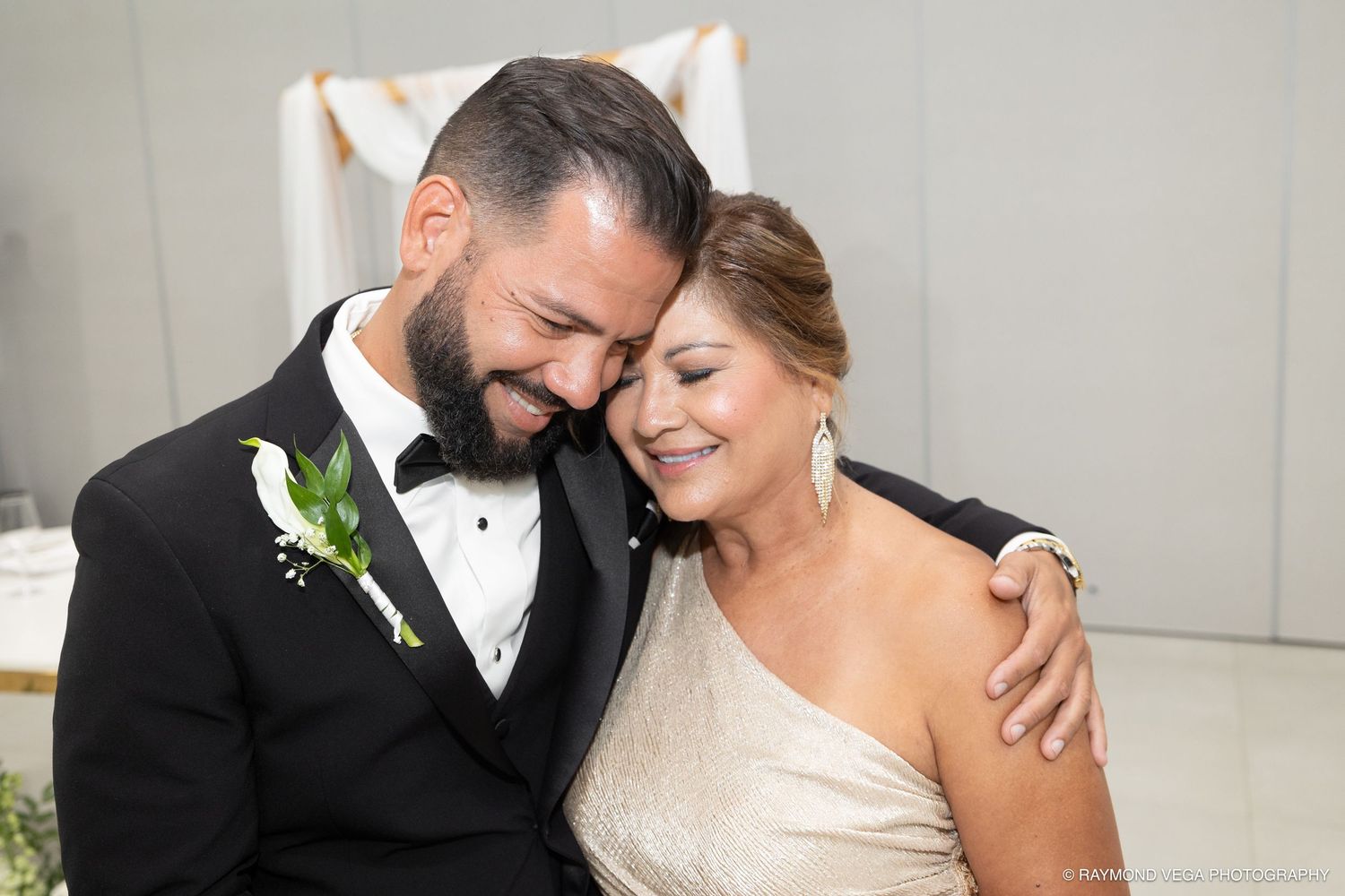 Groom in black tuxedo tenderly embraces smiling bride in gold gown at elegant wedding ceremony.