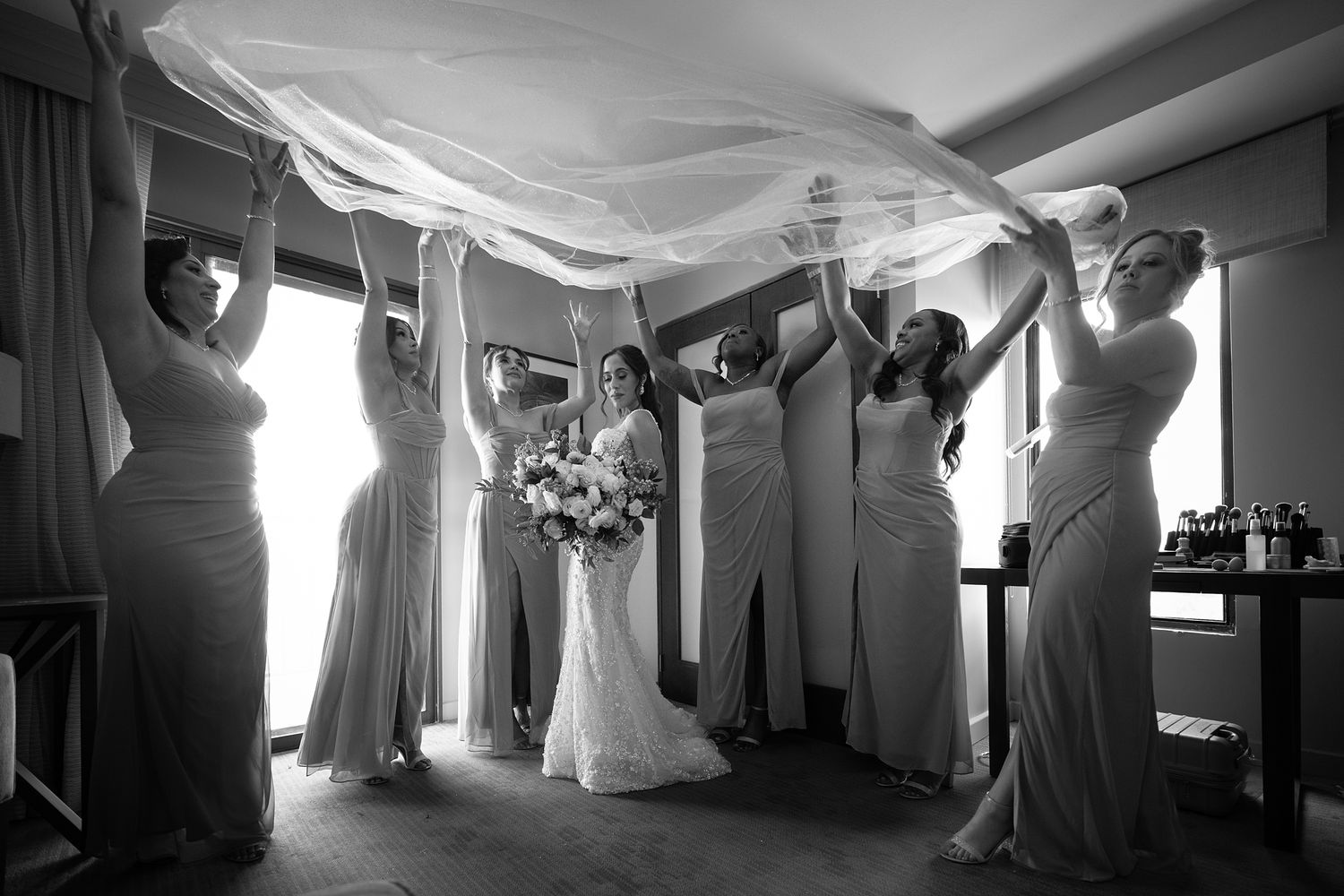 Black and white photo of bride and bridesmaids lifting a flowing veil indoors before the wedding ceremony. Raymond Vega Photography & Cinema