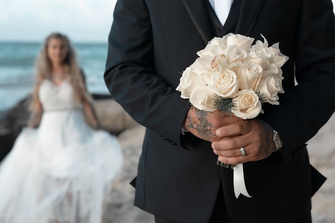 Groom in black suit holds white rose bouquet on beach, tattooed hands, bride in white gown blurred in background.