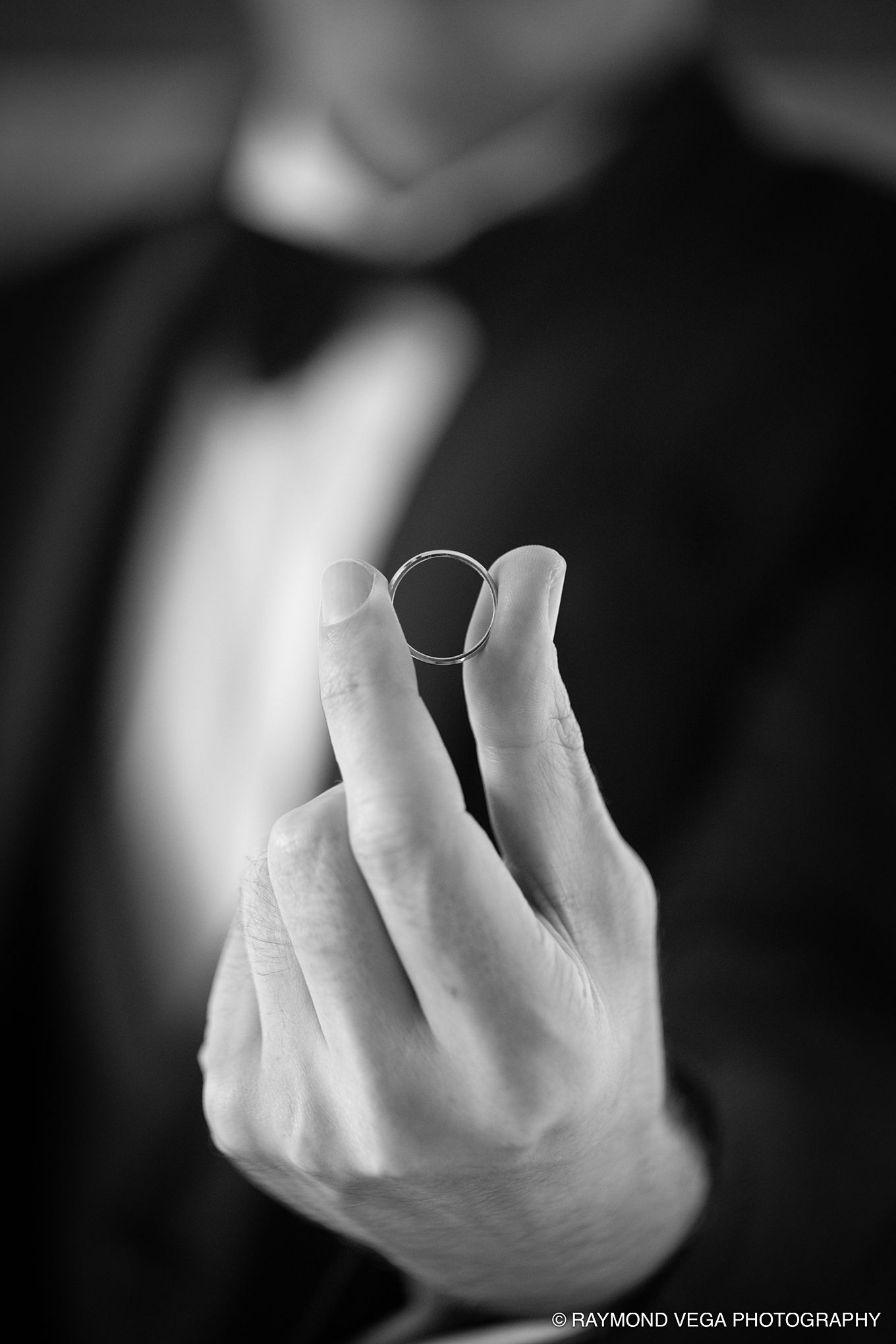 Black and white photo of a groom in a tuxedo holding a wedding ring between his fingers.