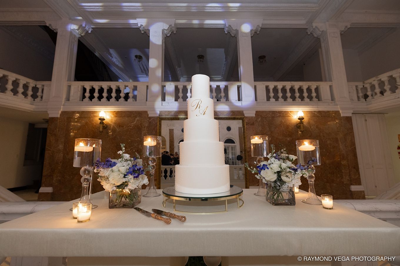 Elegant white multi-tier wedding cake displayed on a table in a grand ballroom with marble columns and candlelight.
