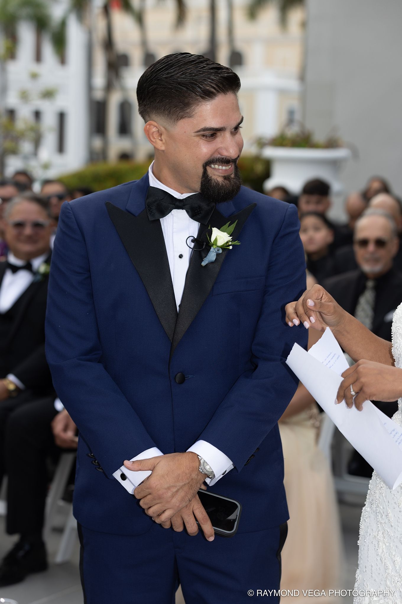 Groom in navy blue tuxedo smiling during outdoor wedding ceremony, wearing boutonniere and bow tie. Antiguo Casino Puerto Rico