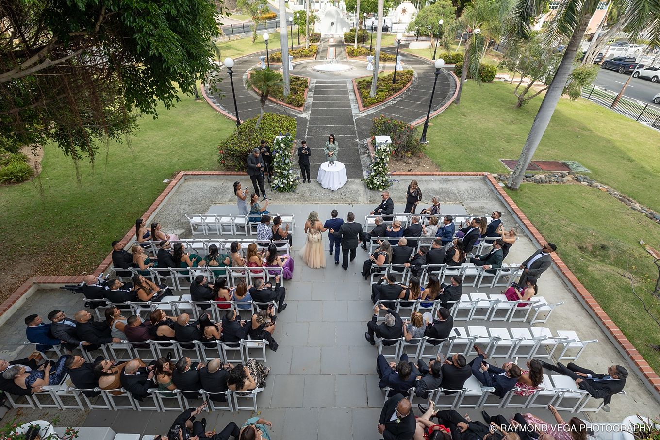 Aerial view of an elegant outdoor wedding ceremony with guests seated in white chairs on a patio surrounded by lush greenery.