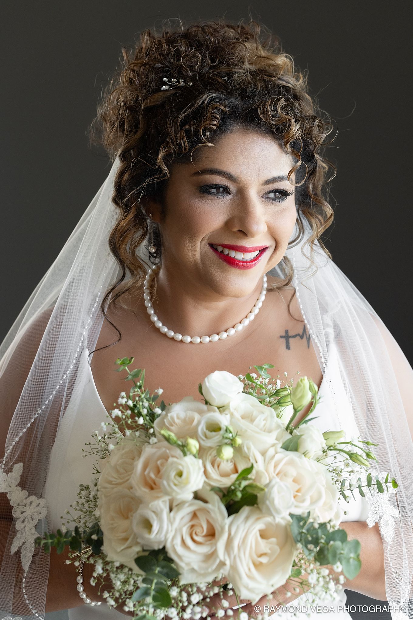 Smiling bride with curly hair, white veil, pearl necklace, red lips, and a bouquet of white roses on her wedding day.