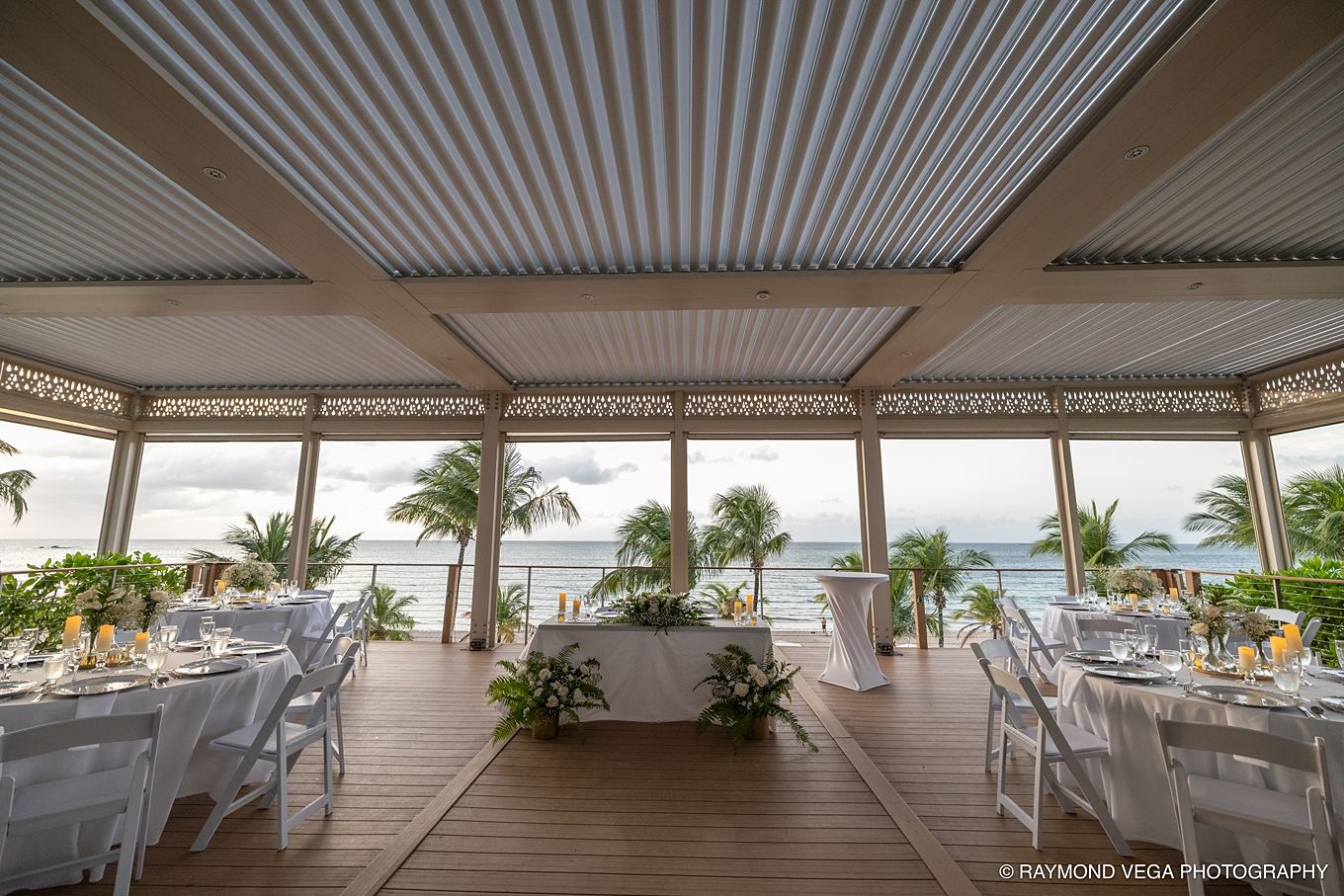 Elegant beachfront wedding venue with white tables, tropical palms, and ocean views under a modern louvered pergola roof. at Courtyard