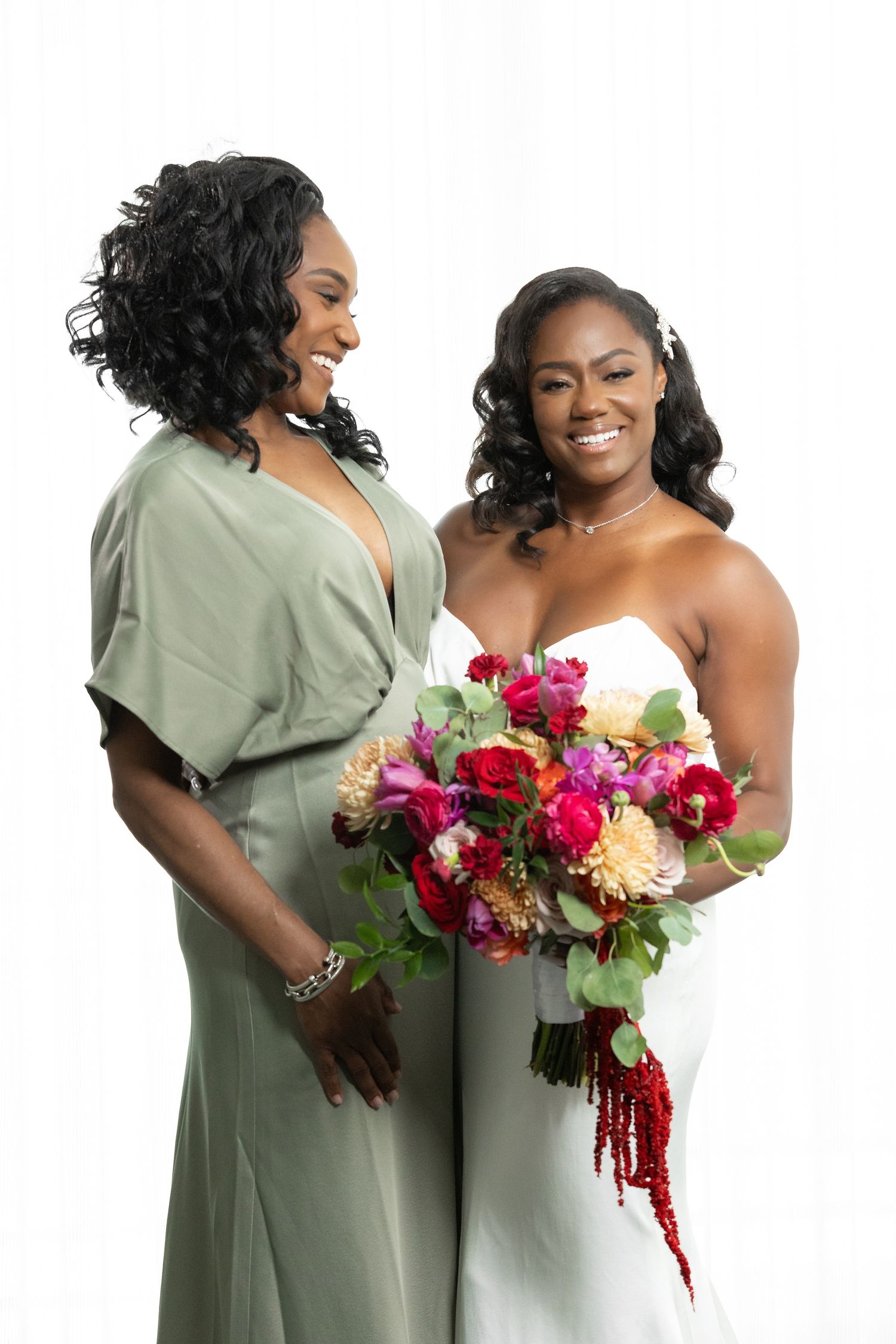 Bride in white strapless gown holds colorful bouquet beside smiling bridesmaid in sage green dress on white background.