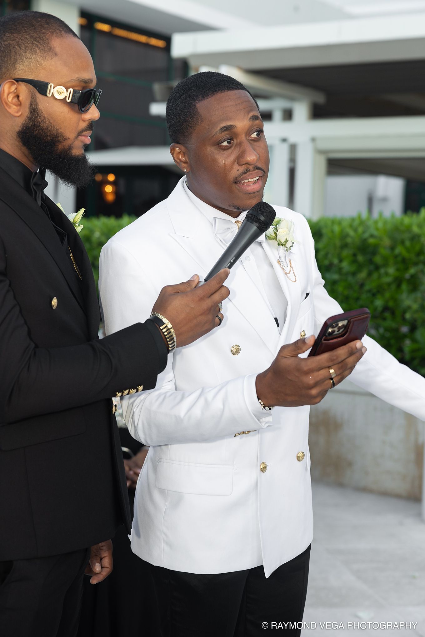 Man in white tuxedo jacket speaks into microphone at formal event while holding phone outdoors.