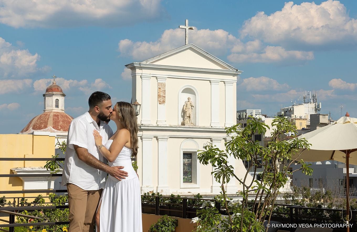 Couple sharing a romantic kiss on a rooftop with a white church and blue sky in the background.Hotel El convento Old San Juan